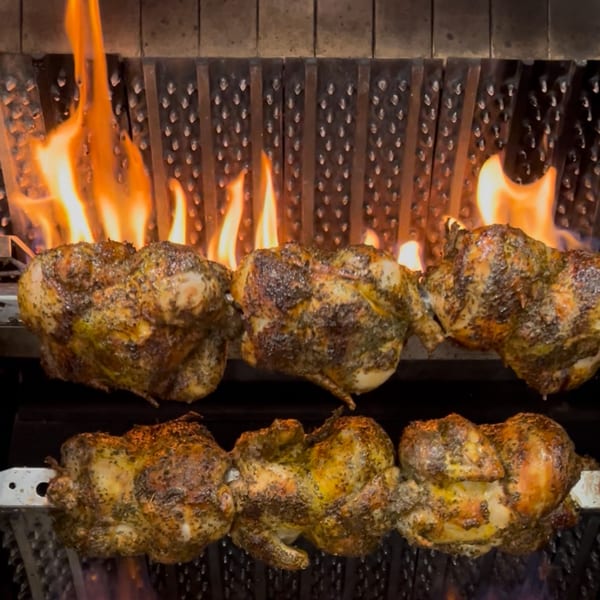 Six seasoned whole chickens rotisserie cooking over an open flame in a dark oven.